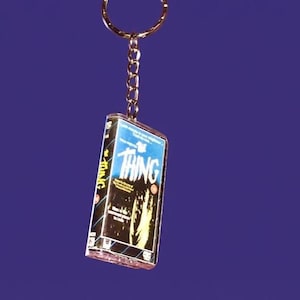 Peut inclure: Porte-clés représentant une mini-cassette VHS du film "The Thing". La cassette a une couverture bleue et noire avec le titre en grandes lettres blanches. Il est suspendu à une chaîne et un anneau dorés.