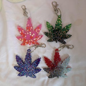 May include: Four leaf-shaped keychains made of resin with metal lobster claw clasps. One is pink with heart-shaped glitter, one is dark green with star-shaped glitter, one is purple with star-shaped glitter, and one is a red, black, and white gradient.