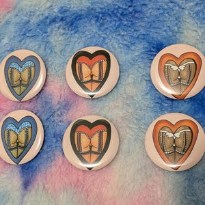 May include: Six circular pin-back badges arranged in two rows. Each badge features a heart-shaped graphic containing an illustration of a person wearing lace-trimmed lingerie and fishnet stockings. The hearts are coloured in blue, red, and orange.