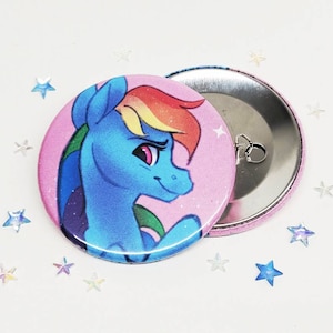 Op de afbeelding: Een ronde knoop met een roze achtergrond en een cartoon afbeelding van een blauw en regenboogkleurig pony met een roze manen en staart. De pony kijkt naar rechts en heeft een serieuze uitdrukking. De knoop heeft een zilveren achterkant met een pin en een lus om hem aan een tas of jas te bevestigen.