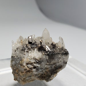 Könnte beinhalten: Eine kleine Gruppe von klaren Quarzkristallen mit einem metallisch grauen Mineral auf einem rauen, grauen Gestein.