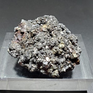 Könnte beinhalten: Dunkles, strukturiertes Mineralexemplar mit einer Mischung aus metallischen Silber-, Schwarz- und rötlich-braunen Farbtönen. Das Mineral liegt auf einem transparenten, rechteckigen Präsentationsständer vor dunklem Hintergrund. Die Oberfläche des Minerals ist rau und uneben.
