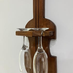 Könnte beinhalten: Ein dunkelbrauner Weinhalter aus Holz in Geigenform. Zwei klare Weingläser hängen an der Halterung, mit zwei kleinen silbernen Untertassen auf dem Regal darüber. Er ist an einer weißen Wand montiert.