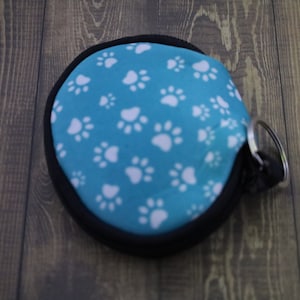 May include: A small, round, blue pouch with a white paw print pattern. The pouch has a black zipper and a silver key ring.