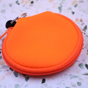 May include: A bright orange round zippered pouch with a silver key ring attached. The pouch is made of a soft, textured fabric.
