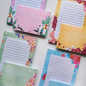 Peut inclure: Ensemble de papeterie à thème floral, comprenant des blocs-notes lignés et des cartes de notes vierges. Les blocs-notes présentent une variété de couleurs, dont le rose, le jaune, le vert et le bleu, avec des motifs floraux d'angle. Les cartes ont un fond de couleur unie avec des motifs floraux d'angle.