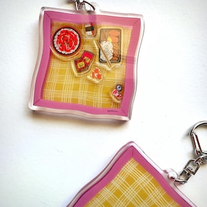 May include: Two clear acrylic keychains with a pink border and a yellow plaid background. Each keychain features miniature illustrations of sushi, bento boxes, and condiments. The keychains have silver-colored metal clasps.