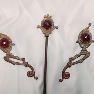 May include: Three antique decorative metal pieces with a vintage aesthetic. The pieces feature ornate designs with a central, faceted, red gemstone. The central piece is a long rod, flanked by two curved, decorative elements.
