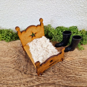 Könnte beinhalten: Ein Miniatur-Holzbett mit einer weißen, flauschigen Decke und zwei schwarzen Gummistiefeln auf einer Holzoberfläche mit grünem Moos.