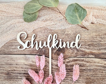 May include: Wooden sign with the word "Schulkind" in cursive script, surrounded by pink dried flowers and green leaves on a wooden background.
