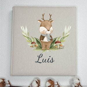 May include: A beige linen photo album with a watercolor illustration of a deer wearing a scarf and a brown jacket. The deer is surrounded by green leaves and red mushrooms. The album has the name "Luis" printed in black cursive lettering.