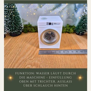 May include: A miniature white washing machine with a silver-colored door and a ruler next to it. The text on the image says "FUNKTION: WASSER L&Auml;UFT DURCH DIE MASCHINE - EINF&Uuml;LLUNG OBEN MIT TRICHTER, AUSLASS &Uuml;BER SCHLAUCH HINTEN."