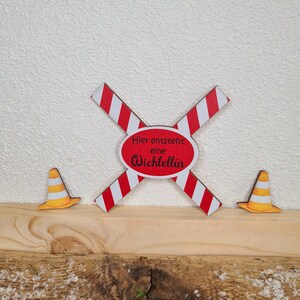 Pu&ograve; includere: Un cartello di legno a strisce rosse e bianche con il testo "Hier entsteht eine Wichtelt&uuml;r" in lettere nere. Due coni di segnalazione gialli sono su entrambi i lati del cartello.