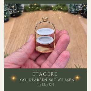 Könnte beinhalten: Ein goldfarbener Miniatur-Servierständer mit zwei Etagen und zwei hellblauen Tellern. Der Ständer hat einen Griff oben und wird in einer Hand gehalten. Der Text auf dem Bild lautet "ETAGERE GOLDFARBEN MIT WEISSEN TELLERN."