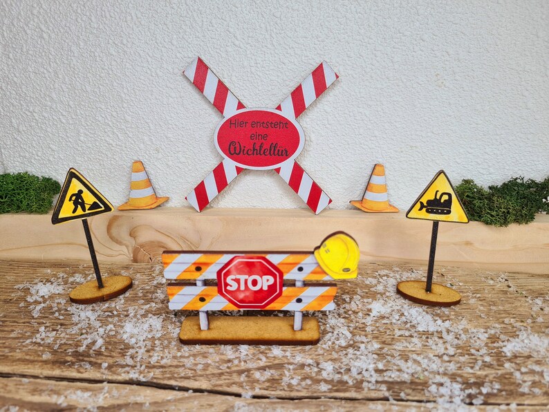 Pu&ograve; includere: Un set di cartelli di cantiere in legno con una barriera a strisce rosse e bianche, un cartello di stop giallo, un cono di cantiere giallo, un cartello giallo con un operaio, un cartello giallo con un bulldozer e un casco giallo. I cartelli indicano "Hier entsteht eine Wichtelt&uuml;r" e "STOP".