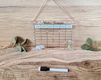 May include: A clear acrylic dry-erase board with a blue header that says "Valentin's Stundenplan". The board is divided into a grid with days of the week labeled across the top and numbers 1 through 7 down the side. There are two dinosaur figures on the board, a green dinosaur on the right and a gray dinosaur on the left. A white dry-erase marker is lying on a wooden surface below the board.