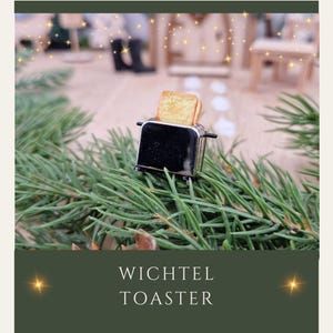 Op de afbeelding: Een miniatuur zwarte en zilveren broodrooster met twee sneetjes goudbruin toast, tegen een achtergrond van groene dennennaalden. De tekst "WICHTEL TOASTER" staat eronder.