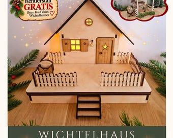 May include: A light-colored wooden miniature house with a peaked roof, a small door with a star cutout, a window with shutters, and a picket fence. The structure features interior lighting, a small set of front steps, and a miniature mailbox.