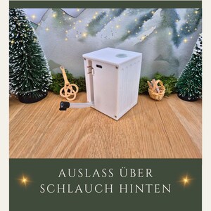 May include: A white, rectangular device with a spout and a small, round opening on top. The device has a ridged side and is positioned on a wooden surface. The text "AUSLASS &Uuml;BER SCHLAUCH HINTEN" is at the bottom of the image.