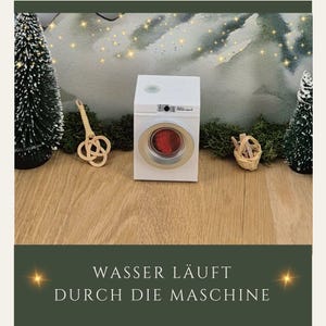 May include: A miniature white washing machine with a red interior, set on a wooden surface. The machine has a silver-colored door frame. The text at the bottom reads "WASSER L&Auml;UFT DURCH DIE MASCHINE". Decorative elements include small Christmas trees and a woven basket.