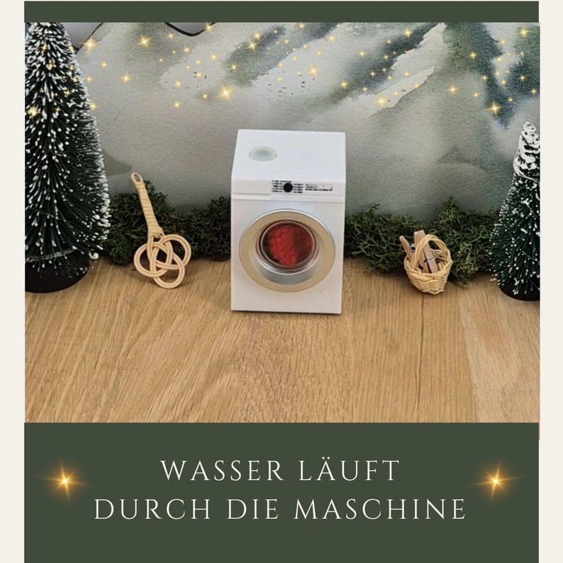 May include: A miniature white washing machine with a red interior, set on a wooden surface. The machine has a silver-colored door frame. The text at the bottom reads "WASSER L&Auml;UFT DURCH DIE MASCHINE". Decorative elements include small Christmas trees and a woven basket.