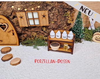 Könnte beinhalten: Ein Miniatur-Holzregal mit vier weißen Porzellanbehältern, die mit "Zucker", "Tee", "Reis" und "Mehl" beschriftet sind. Das Regal ist mit einem kleinen Holzsieb und einem Schneebesen verziert. Das Regal ist vor dem Hintergrund einer Holztür und einer Schneelandschaft platziert.