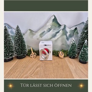 May include: A miniature white washing machine with an open red door is the focal point, surrounded by small green Christmas trees and a mountain backdrop. The text "T&Uuml;R L&Auml;SST SICH &Ouml;FFNEN" is at the bottom.
