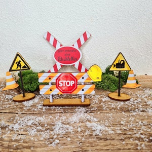 Pu&ograve; includere: Un set di decorazioni per feste a tema edile in legno con una croce a strisce rosse e bianche, una barriera a strisce gialle e nere con un cartello di stop rosso, un cartello di cantiere giallo con una sagoma nera di un operaio, un cartello di cantiere giallo con una sagoma nera di un bulldozer e un casco giallo. I cartelli sono su supporti in legno.