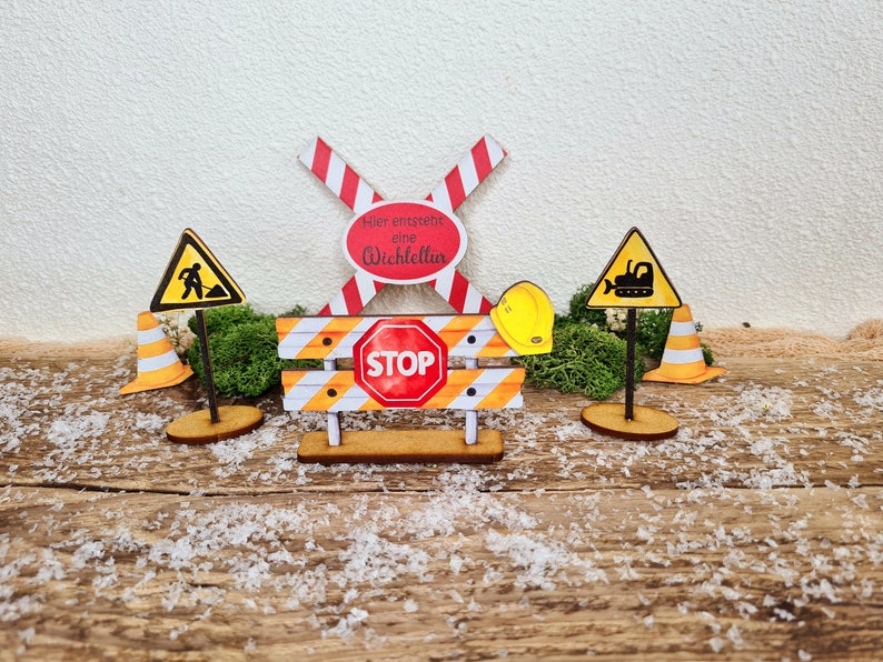 Pu&ograve; includere: Un set di decorazioni per feste a tema edile in legno con una croce a strisce rosse e bianche, una barriera a strisce gialle e nere con un cartello di stop rosso, un cartello di cantiere giallo con una sagoma nera di un operaio, un cartello di cantiere giallo con una sagoma nera di un bulldozer e un casco giallo. I cartelli sono su supporti in legno.