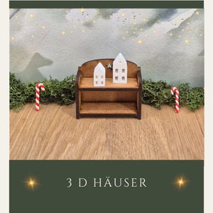 Könnte beinhalten: Ein Miniatur-Holzregal mit zwei kleinen weißen Häusern und zwei Zuckerstangen. Die Häuser haben ausgeschnittene Fenster und einen Stern. Der Text "3 D HÄUSER" befindet sich am unteren Rand des Bildes.