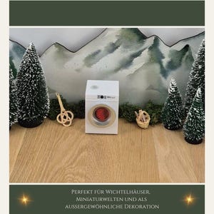 May include: A miniature white washing machine with a red interior, set against a backdrop of miniature evergreen trees and a mountain scene. A small wooden decorative item and a tiny basket are also present. The image includes text at the bottom.