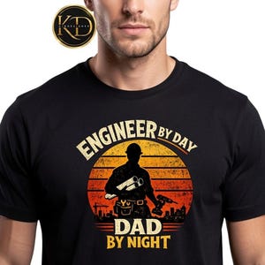 May include: Black t-shirt with a graphic design featuring an engineer silhouette holding blueprints and a drill, set against a sunset backdrop. Text reads "ENGINEER BY DAY, DAD BY NIGHT."