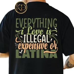 May include: A black t-shirt featuring a graphic design on the back with the text "EVERYTHING i Love is ILLEGAL expensive or LATINA" in shades of olive green, cream, and peach, accented with decorative swirls and a circular logo in the top corner.