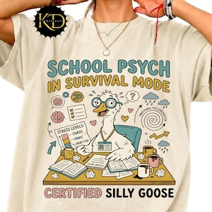 Könnte beinhalten: Cremefarbenes T-Shirt mit einer Grafik, die eine Comic-Gans mit Brille zeigt, die an einem Schreibtisch mit Büchern und Tassen sitzt. Der Text lautet "SCHOOL PSYCH IN SURVIVAL MODE" und "CERTIFIED SILLY GOOSE". Das Design enthält Stresslevel-Indikatoren und andere skurrile Elemente.