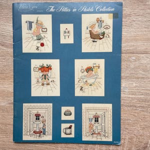 May include: A blue booklet titled "The Potties in Pastels Collection" by Alma Lynne. The booklet features cross-stitch patterns of bathroom scenes with cartoon characters. The cover has a blue background and the images are in pastel colors.