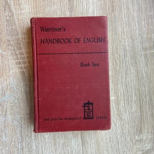May include: A red hardcover textbook titled Warriner's Handbook of English Book Two from The English Workshop Series. The front cover features black text and a small black graphic of a printing press centered near the bottom of the book.