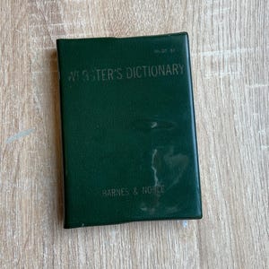 May include: A dark green Webster's Dictionary book from Barnes & Noble. The book has a rectangular shape and is lying flat on a light wood surface. The title is printed in white letters.