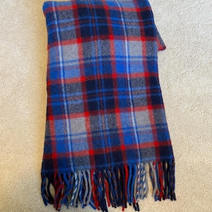 May include: A folded plaid scarf in shades of blue, red, and gray. The scarf features a classic tartan pattern with fringed edges. The scarf is made of a soft material, perfect for cold weather.