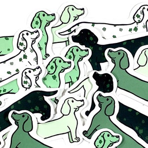 May include: A collection of green, black, and white dachshund stickers with a shamrock pattern. The stickers are arranged in a pile, with the heads of the dachshunds facing different directions.