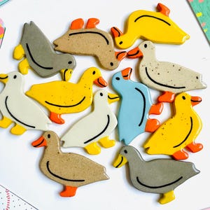 May include: A collection of duck-shaped ceramic magnets in various colors, including yellow, gray, white, and blue, with orange beaks and feet. The magnets are arranged on a white surface, surrounded by colorful cards with floral and abstract designs.