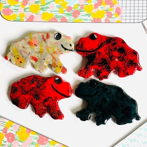 Pode incluir: Quatro estatuetas de sapo de cerâmica. Um sapo é branco com manchas marrons, laranja e amarelas. Dois sapos são vermelhos com manchas pretas. Um sapo é preto com manchas brancas. Os sapos estão sobre uma superfície branca.