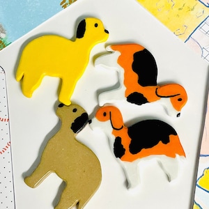 May include: Four ceramic dog figurines in different colors and poses. The dogs are yellow, brown, black and white, and orange and white.