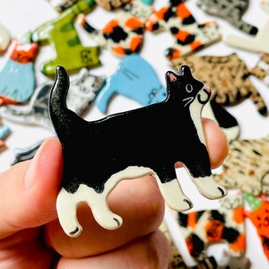 May include: A ceramic cat-shaped pin featuring a black body with white markings on the face, chest, and paws. The cat is depicted in a standing position with its tail curved upward. The surface has a glossy finish, and the edges are smooth.