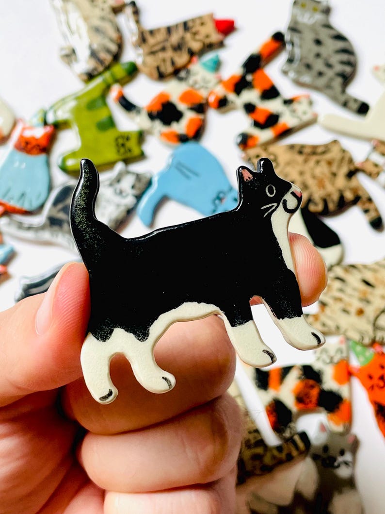 May include: A ceramic cat-shaped pin featuring a black body with white markings on the face, chest, and paws. The cat is depicted in a standing position with its tail curved upward. The surface has a glossy finish, and the edges are smooth.
