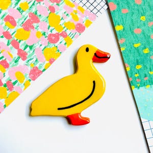 May include: A yellow duck-shaped pin with a black curved line and red feet and beak. The pin is on a white surface with colorful patterned paper in the background. The duck pin is a fun, decorative accessory.