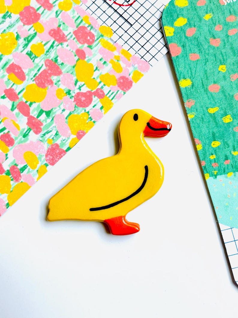 May include: A yellow duck-shaped pin with a black curved line and red feet and beak. The pin is on a white surface with colorful patterned paper in the background. The duck pin is a fun, decorative accessory.