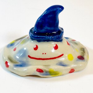May include: A ceramic figurine of a smiling creature with a blue hat. The creature has a white body with red and yellow spots.