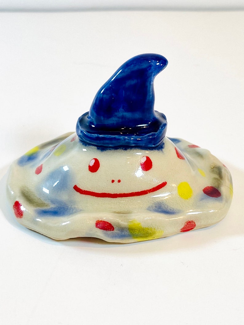 May include: A ceramic figurine of a smiling creature with a blue hat. The creature has a white body with red and yellow spots.