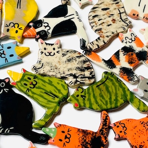 May include: A collection of small, flat, ceramic cat-shaped ornaments in various colors and patterns, including solid black, orange, green, and multi-colored calico designs. Several of the cats feature small, pointed party hats on their heads.