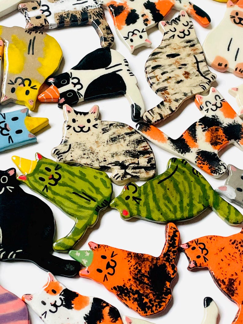 May include: A collection of small, flat, ceramic cat-shaped ornaments in various colors and patterns, including solid black, orange, green, and multi-colored calico designs. Several of the cats feature small, pointed party hats on their heads.